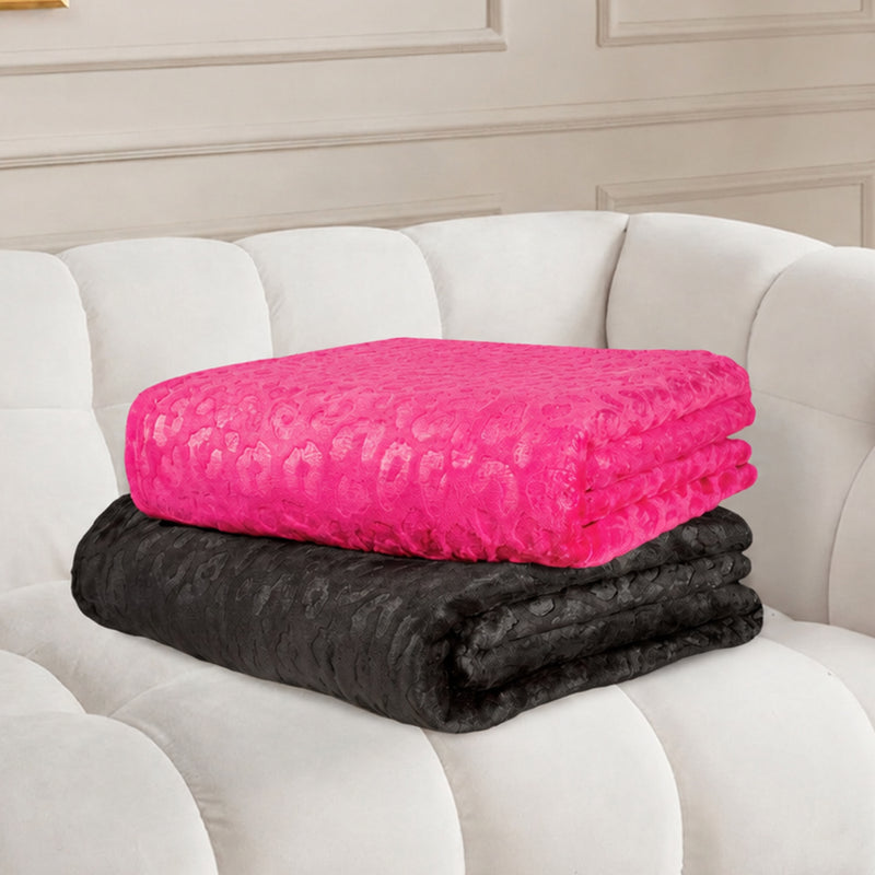 Two folded blankets, one bright pink and one black, with a subtle animal print texture, are stacked on a plush white couch against a cream-colored decorative wall. The vibrant pink throw adds a pop of color and style, while the black Embossed Plush Oversized Throw Blanket features an embossed finish for extra softness and sophistication.