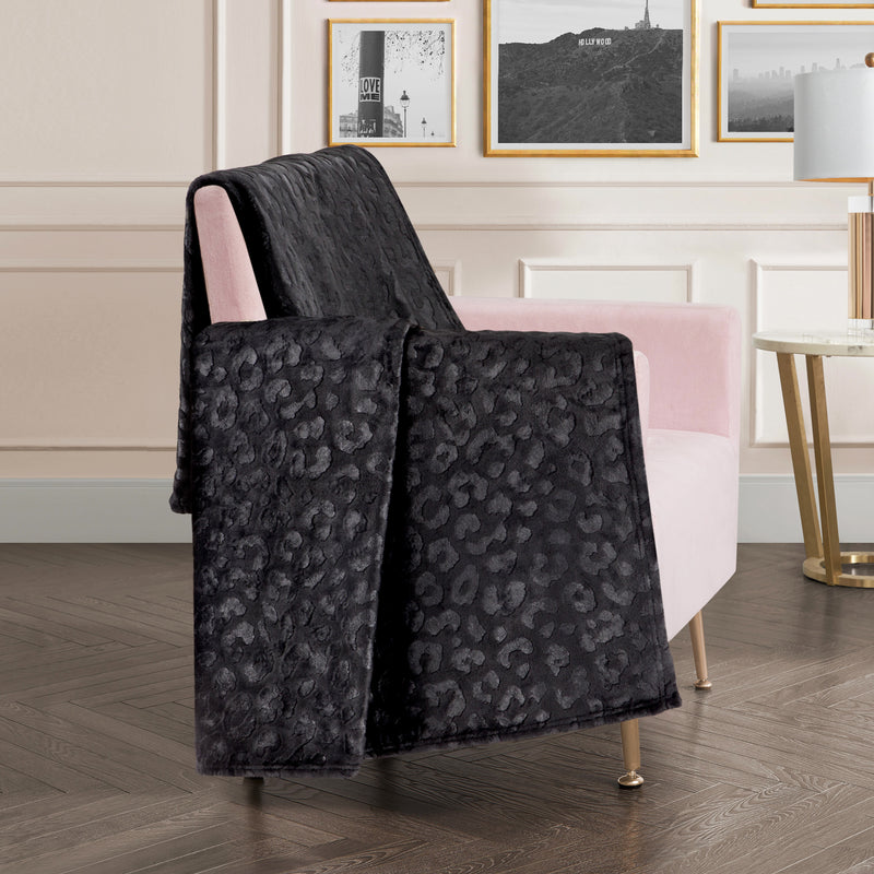 An Embossed Plush Oversized Throw Blanket with a black leopard print is draped over a light pink armchair in a modern living room featuring wood flooring, framed art, and a round side table. The velvety soft blanket adds both style and comfort to the chic space.