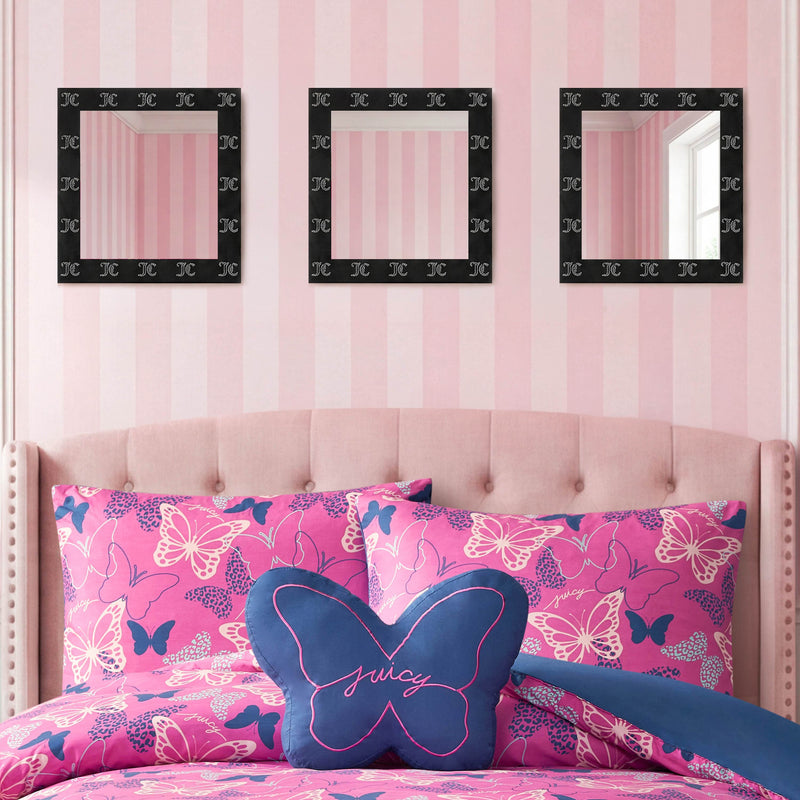 A pink and white striped bedroom wall features the Velour Rhinestone Logo 3-Piece Decorative Mirror Set, with black frames and ready-to-hang design, displayed above a tufted pink headboard. The bed is adorned with pink butterfly-patterned bedding and a blue butterfly-shaped pillow that says "Juicy.