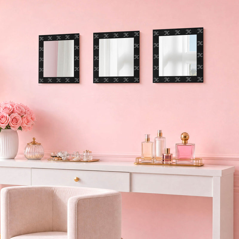 A white vanity topped with perfume bottles and pink flowers is positioned against a pink wall, decorated above by the Velour Rhinestone Logo 3-Piece Decorative Mirror Set, which features three square, ready-to-hang mirrors. In front of the vanity sits a light-colored chair with a velour-look frame.