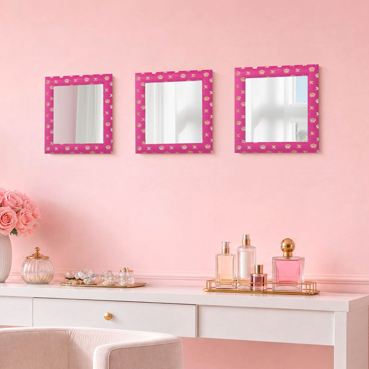 A vanity adorned with a white desk, pink perfume bottles, pink roses in a vase, and the Velour Crown Foil Print Hot Pink with Gold Foil 3-Piece 12 in x 12 in Decorative Mirror Set against a pale pink wall creates an elegant and charming atmosphere. Each mirror is ready-to-hang, allowing you to effortlessly enhance your room with graceful style.