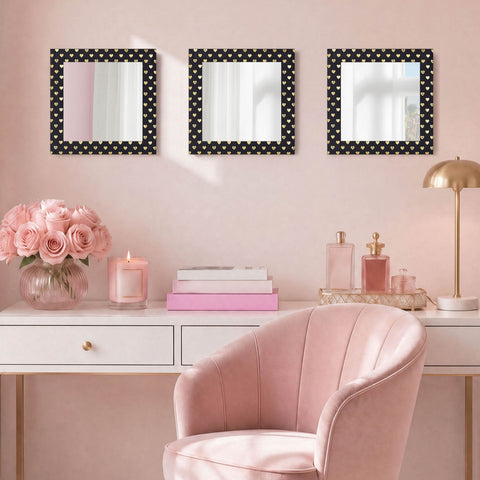 A pink vanity area with a velvet chair, white desk, flowers, candles, perfume bottles, and the Velour Heart Foil Print 3-Piece Decorative Mirror Set featuring black and gold heart patterns as stylish wall décor against the pink wall.