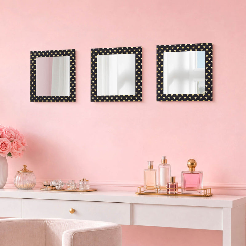 A vanity with perfume bottles sits against a pink wall, above which hangs the Velour Heart Foil Print 3-Piece Decorative Mirror Set—three square mirrors with black frames adorned with small white hearts. Pink flowers and glass containers are arranged on the vanity, enhancing the charming wall décor.