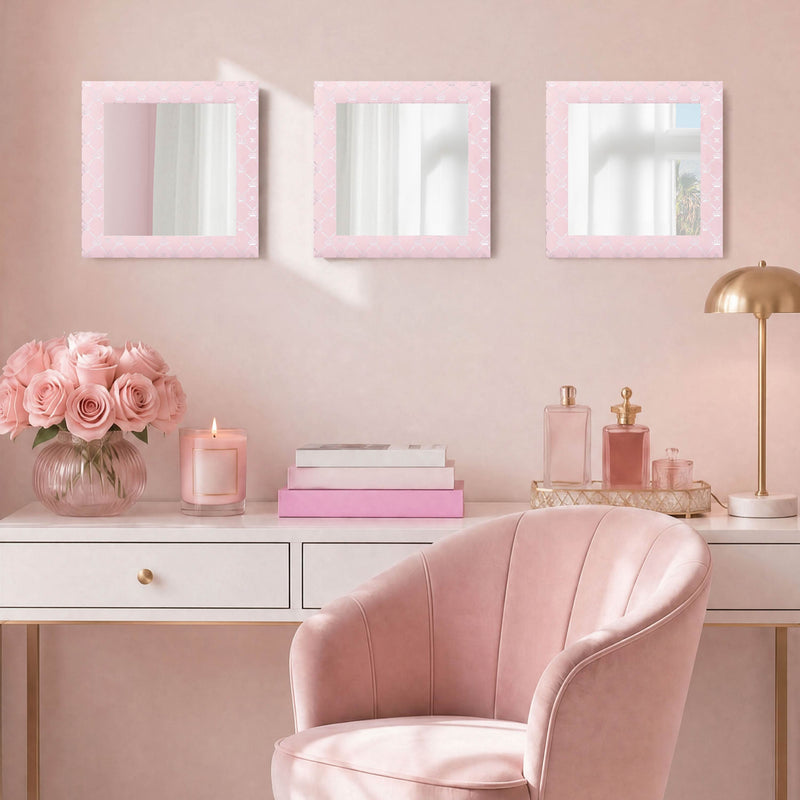 A blush pink vanity with a velvet chair, pink roses in a vase, a candle, books, and a gold lamp is set against a pink wall adorned above with the Velour Lattice Crown Foil Print 3-Piece Decorative Mirror Set as stylish wall decor.