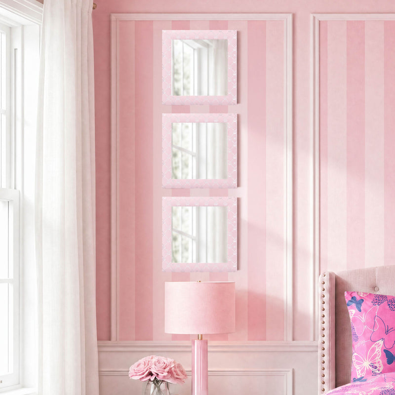 A pink bedroom wall is adorned with the Velour Lattice Crown Foil Print 3-Piece Decorative Mirror Set, featuring three rectangular mirrors stacked vertically. This elegant arrangement is complemented by a pink lamp, pink roses, and a window dressed with white curtains, while part of a bed with a butterfly-patterned pillow adds an extra touch of charm to the thoughtfully styled space.