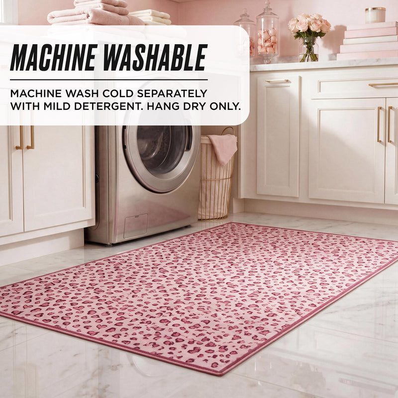 A Finley Washable Rug, 2′ x 4′, with a pink leopard print pattern on a laundry room floor in front of a washing machine, overlaid with text: MACHINE WASHABLE. MACHINE WASH COLD SEPARATELY WITH MILD DETERGENT. HANG DRY ONLY.