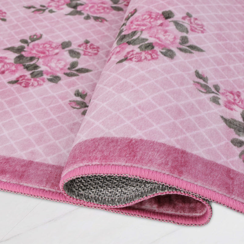 The Finley Washable Rug, 2′ x 4′ is an ultra-soft pink rug with a white lattice pattern and a grey floral design, shown folded to reveal its textured backing.