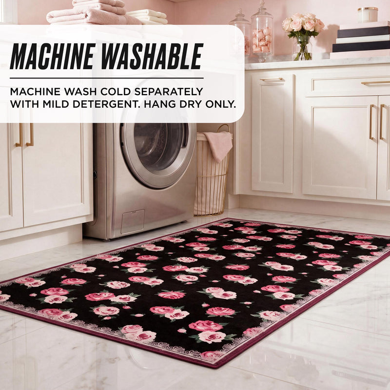 Finley Washable Rug, 2′ x 4′, a rectangular black machine-washable rug with a pink rose floral pattern and a decorative border on a white tiled laundry room floor, featuring a text overlay with care instructions.