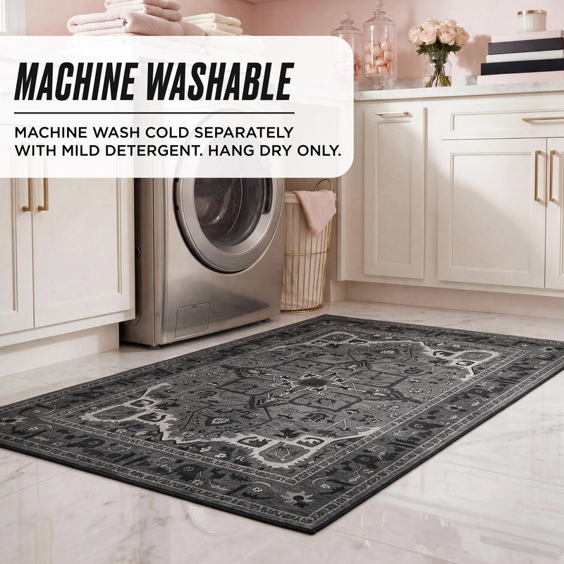 Finley Washable Rug, 2′ x 4′, an ultra-soft, rectangular area rug with a dark gray and black intricate traditional pattern on a white tiled laundry room floor in front of a washing machine.