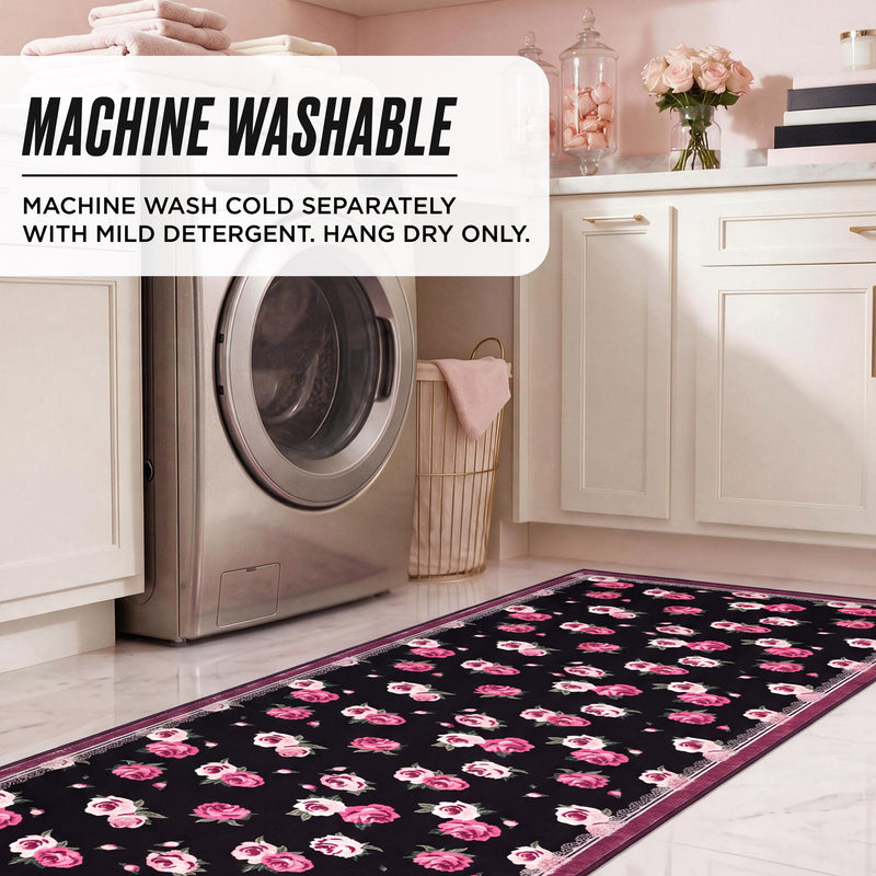 The Finley Washable Rug, 2′ x 6′, is an ultra-soft runner with a black background and pink floral pattern on a tiled floor in front of a washing machine, featuring instructions to wash cold, use mild detergent, and hang dry only.