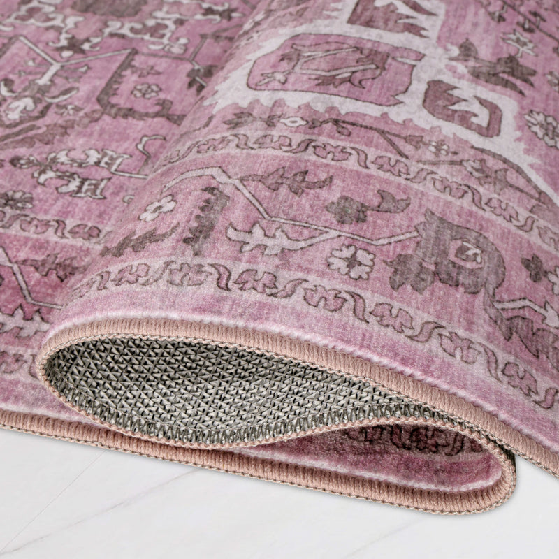 A corner of a dusty pink Finley Washable Rug, 2′ x 4′ with a faded traditional pattern, folded to show the underside with a textured grip backing.