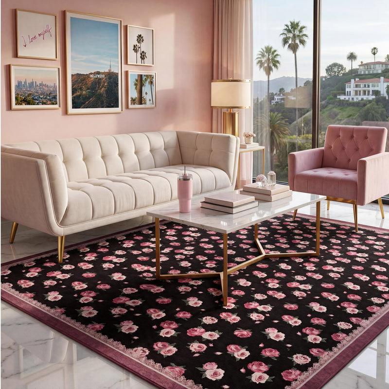 A black Finley Washable Rug, 5′ x 7′, with a repeating pink rose pattern and a dark red border, positioned under a marble coffee table in a living room.