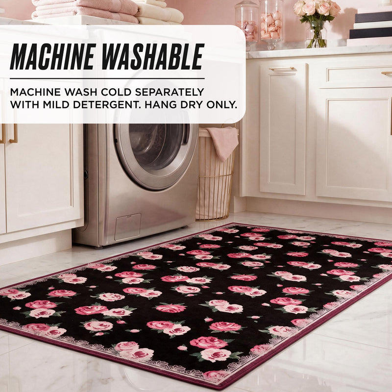 A rectangular black Juicy Couture Finley Washable Rug, 5′ x 7′, with a pink rose floral pattern on a white floor in front of a washing machine; text reads: Machine washable. Machine wash cold separately with mild detergent. Hang dry only.