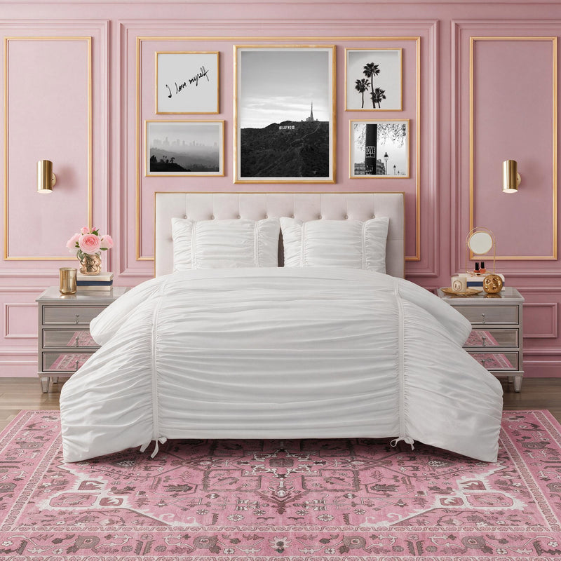 The Finley Washable Rug, 5′ x 7′, is an ultra-soft, machine-washable rectangular area rug featuring a traditional floral pattern in shades of pink and white, placed under a bed in a pink bedroom.