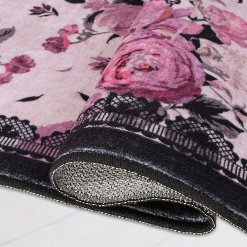 The Finley Washable Rug, 2′ x 6′, is an ultra-soft runner with a faded pink floral pattern and black lace-style border, shown with one end folded over to reveal the textured gray backing.
