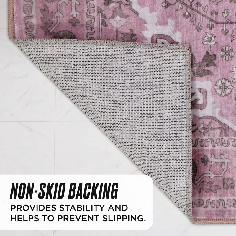 A corner of the pink patterned Finley Washable Rug, 8′ x 10′ is folded back to reveal its textured non-skid backing, with text overlay stating that the backing provides stability and helps prevent slipping.