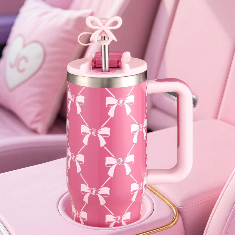 A Jetsetter Tumbler with Handle, Straw & Topper, 30 oz by Juicy Couture, in a large pink design with white bow patterns, sits in a car cupholder. The stainless steel tumbler features a handle, pink lid, and straw topped with a decorative pink bow. Pink car seats are visible in the background.