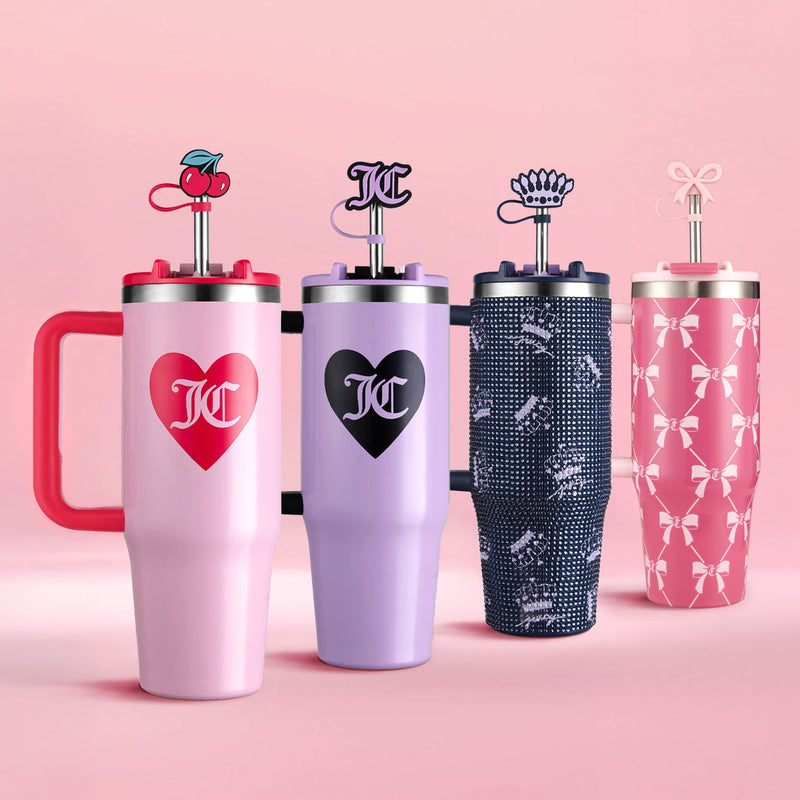 Four vibrant Juicy Couture Jetsetter Tumblers with Handles, Straws & Toppers (30 oz) are displayed against a pink background, each featuring unique patterns—hearts, cherries, bows, and crowns—along with matching lids and decorative stirrers. The initials “JC” are printed on the front of every travel cup.