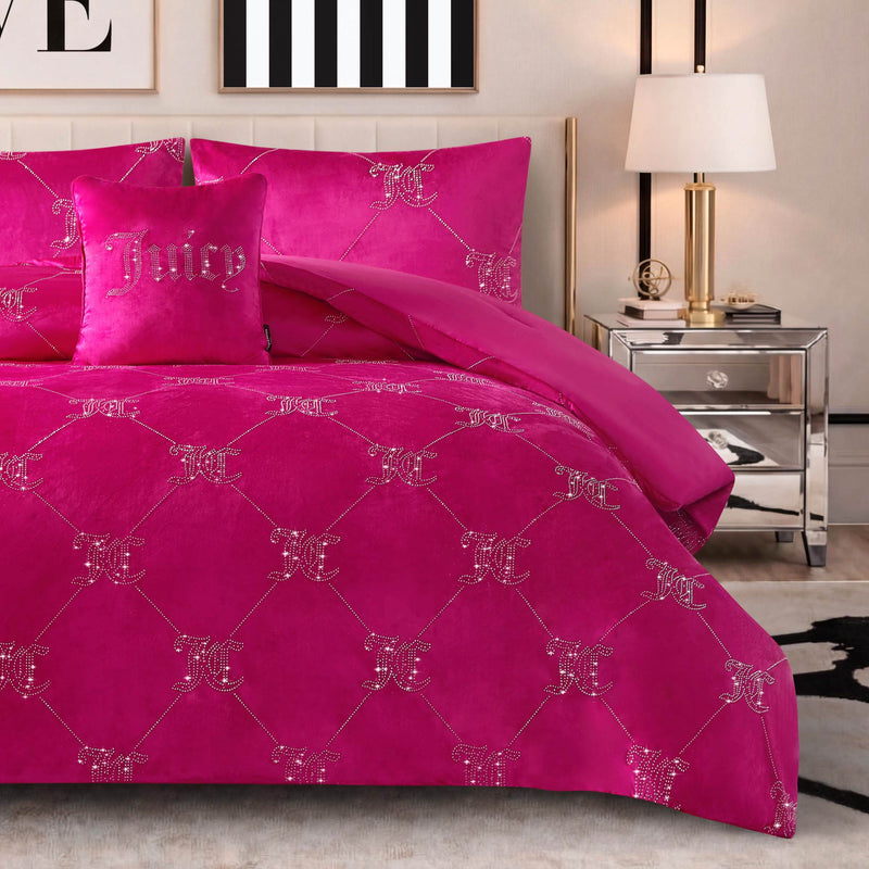 A bed is dressed in the Velvet Rhinestone Logo Link Bling Comforter Set in hot pink, showcasing sparkling rhinestone JC and Juicy logos, and stands beside a mirrored nightstand topped with a lamp; above the bed, abstract wall art completes the look.