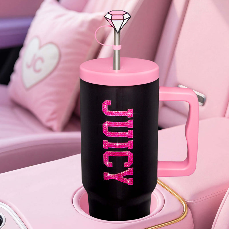 The GoGlam Hydration Bling Stainless Steel Tumbler with Handle and Straw with Topper, 40 oz, in black and pink, sits securely in a car cupholder. Its lid features a reusable straw topped with a diamond-shaped decoration, while vibrant pink car seats and a heart pattern are visible in the background. The tumbler showcases the word “JUICY” in glittery pink letters for extra sparkle.