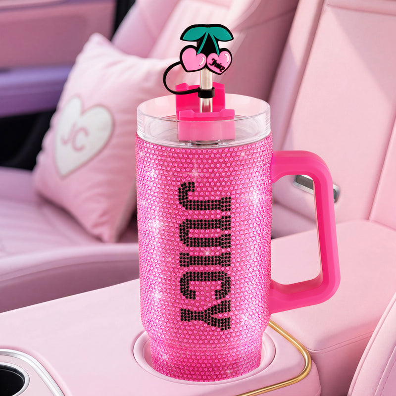 A BlingSip Rhinestone Tumbler with Handle, adorned in sparkling pink rhinestones and labeled JUICY on the side, rests in a car cup holder. This insulated stainless steel tumbler features a lid topped with a cherry-shaped decoration and includes a straw. A heart-shaped pillow can be seen on the seat.