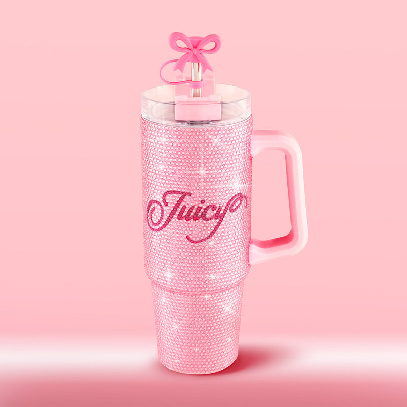 A pink BlingSip Rhinestone Tumbler with a handle and straw sits on a pink background. The insulated stainless steel cup is covered in rhinestones, with the word “Juicy” written in script across the front, and the straw is topped with a bow-shaped topper.