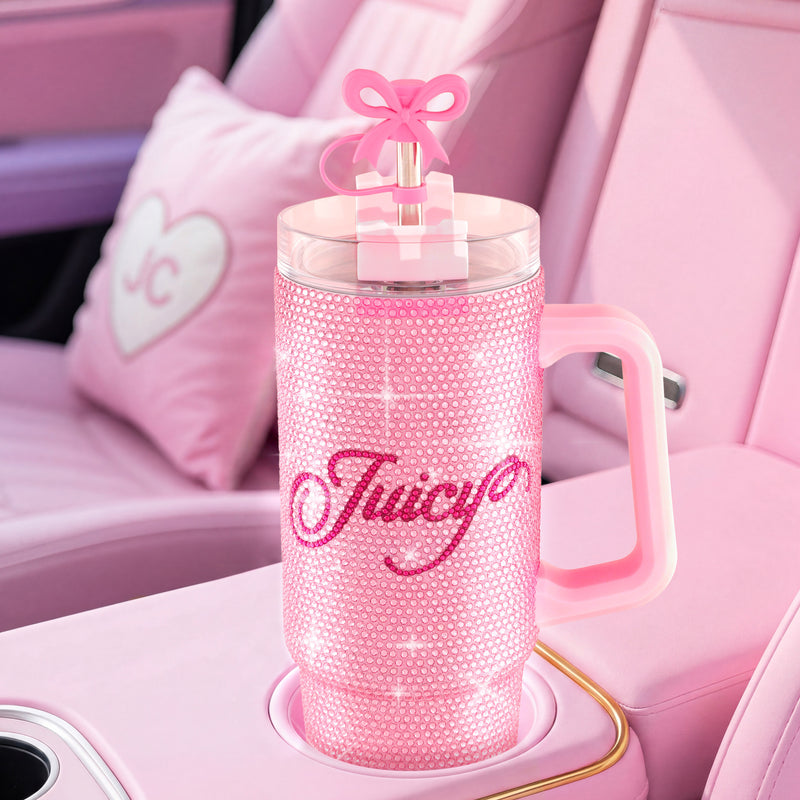 A large pink BlingSip Rhinestone Tumbler with Handle, Straw, and Topper sits in a car cupholder. Its insulated stainless steel interior keeps drinks perfectly chilled, while the sparkling rhinestone exterior stands out. The car’s interior is pink, accented by a heart-shaped pillow and a matching pink straw topped with a bow on the tumbler’s lid.