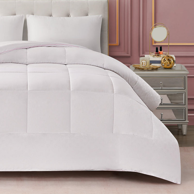 A neatly made bed featuring the white Embossed J Down Alternative Comforter, complemented by a white pillow and a beige tufted headboard, sits beside a mirrored nightstand adorned with a mirror, candle, books, and decorative items—all set against a pink paneled wall.