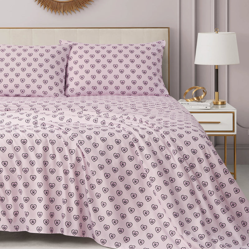 A neatly made bed with a luxury bedding feel is dressed in the Printed Microfiber Sheet Set by Juicy Couture, featuring pink sheets and pillowcases adorned with an outlined heart pattern. The bed sits next to a white and gold nightstand topped with a lamp, books, and decorative objects in a softly lit bedroom.