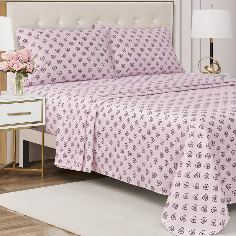 A neatly made bed with a pale pink heart-patterned Printed Microfiber Sheet Set and matching pillowcases, beside a white nightstand adorned with pink flowers and a lamp, features a beige tufted headboard in a softly lit bedroom, exuding an air of luxury bedding.