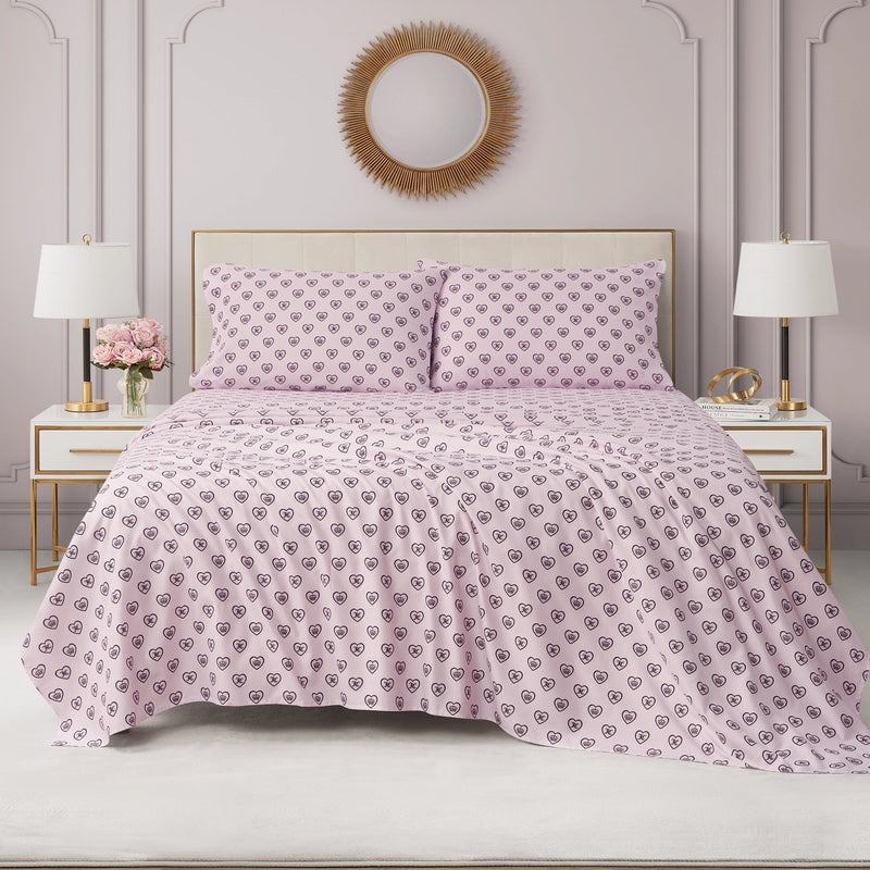 A neatly made bed featuring a pink and purple patterned bedspread, two matching pillows, white nightstands with lamps, flowers, and a round sunburst mirror above the headboard creates an inviting retreat. The look is completed with the Printed Microfiber Sheet Set, which adds a touch of luxury bedding to the space.