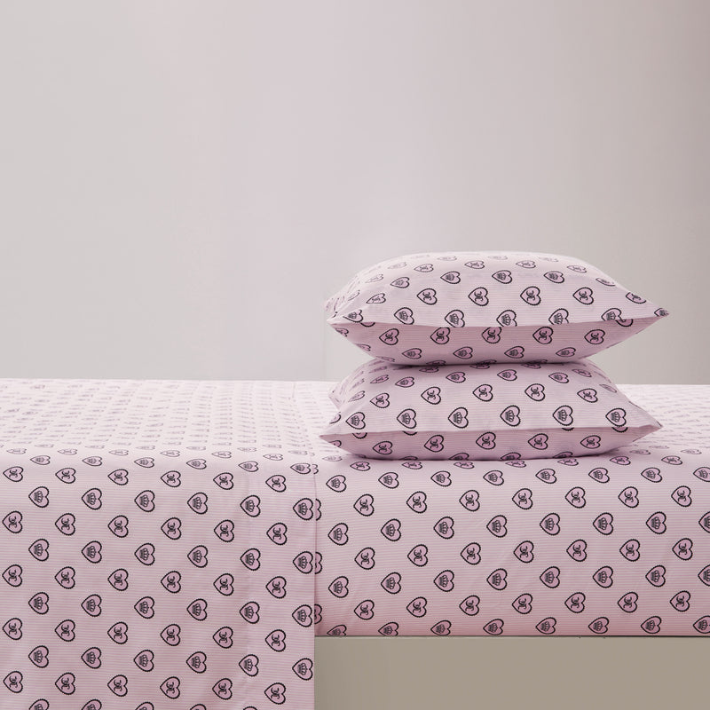 Two stacked pillows and a neatly spread sheet, all in light pink with a repeating pattern of small, outlined black hearts—this Printed Microfiber Sheet Set brings a playful yet elegant touch to any bedroom. The background is plain and light-colored, perfectly highlighting the sheet set’s charming design.