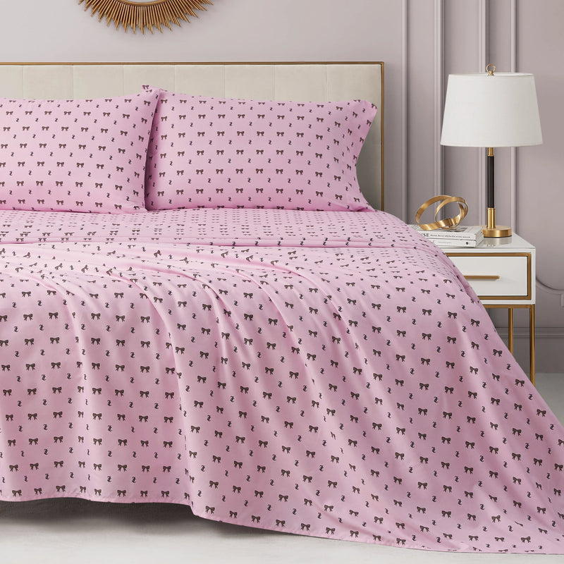 A neatly made bed with a pink Printed Microfiber Sheet Set and matching pillowcases featuring a small black cat pattern, next to a white nightstand with a lamp and gold decor in a modern, softly lit bedroom, creates an inviting touch of luxury bedding.