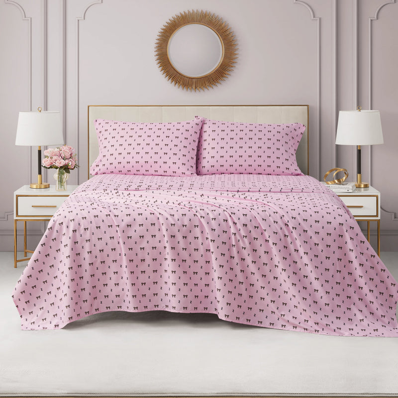 A neatly made bed dressed in a Printed Microfiber Sheet Set with pink sheets and pillowcases featuring a small black pattern is flanked by two white nightstands topped with lamps, flowers, and decor, all set against a pale mauve wall adorned with a round sunburst mirror.