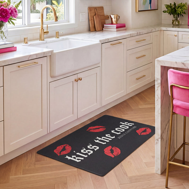 Modern kitchen with white cabinets, gold handles, and a farmhouse sink. A Kitchen Wellness Mat in black featuring red lips and the words “Kiss the Cook” offers anti-fatigue support on the floor. A pink velvet chair is next to the counter.