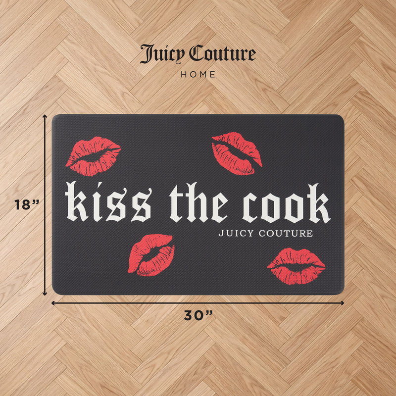 The Kitchen Wellness Mat features a black design with “kiss the cook JUICY COUTURE” in bold white Gothic lettering and four red lips accents. Measuring 18 inches by 30 inches, this mat lies on a wood floor, delivering stylish anti-fatigue support while you cook. “Juicy Couture Home” appears above the mat.