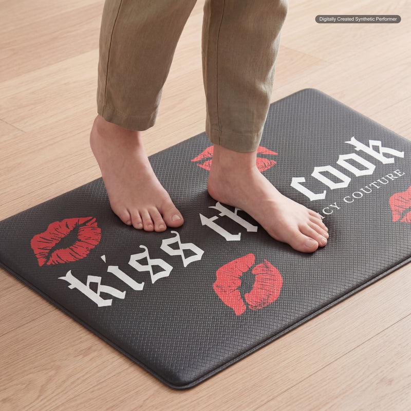 Bare feet stand on the Kitchen Wellness Mat, featuring red lipstick prints and the phrase “kiss the cook” in bold white letters. Placed on a light wood floor, this mat brings both comfort and style to your kitchen space.