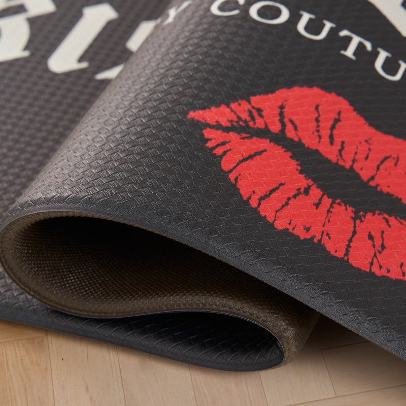 A close-up of the Kitchen Wellness Mat, rolled up in black with a red lipstick print and white text, displayed on a light wooden floor, offering anti-fatigue support for your exercise or kitchen routine.