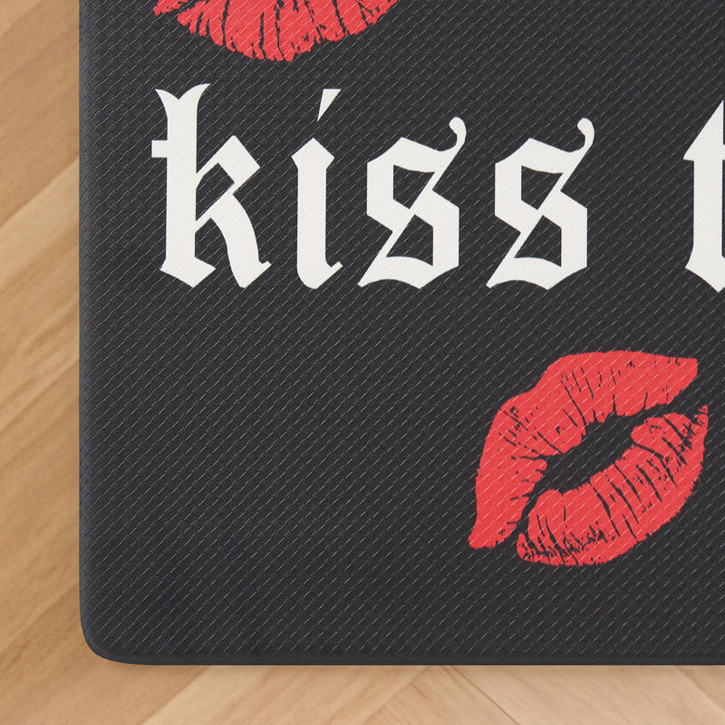 Introducing the Kitchen Wellness Mat—featuring a black textured design with bold white Gothic “kiss” lettering and two vibrant red lipstick lip prints, one above and one below the text. Perfectly placed on a wooden floor, this mat is designed for anti-fatigue support, delivering both comfort and unique personality to your kitchen space.
