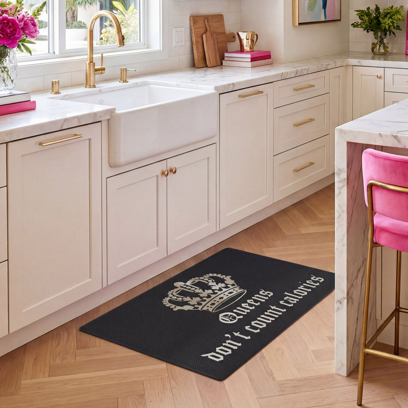 A stylish kitchen features white cabinets with gold handles, a farmhouse sink, and a chic Kitchen Wellness Mat on the floor that reads “Queens don’t count calories” in white alongside a crown graphic. A pink barstool sits beside a marble-topped island, completing the contemporary look.