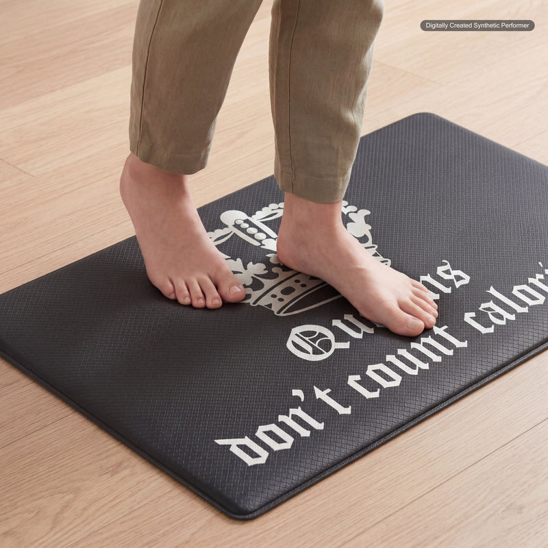 A person stands barefoot on a Kitchen Wellness Mat featuring a white crown graphic and partial text reading “don’t count calo...”. This cushioned anti-fatigue mat is placed on a light wooden floor.