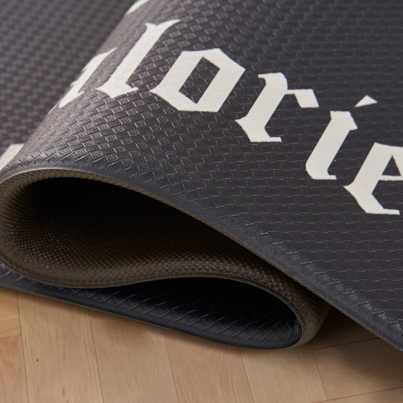 A close-up of a partially rolled-up black Kitchen Wellness Mat with a textured surface and large white letters, resting on a light wooden floor.