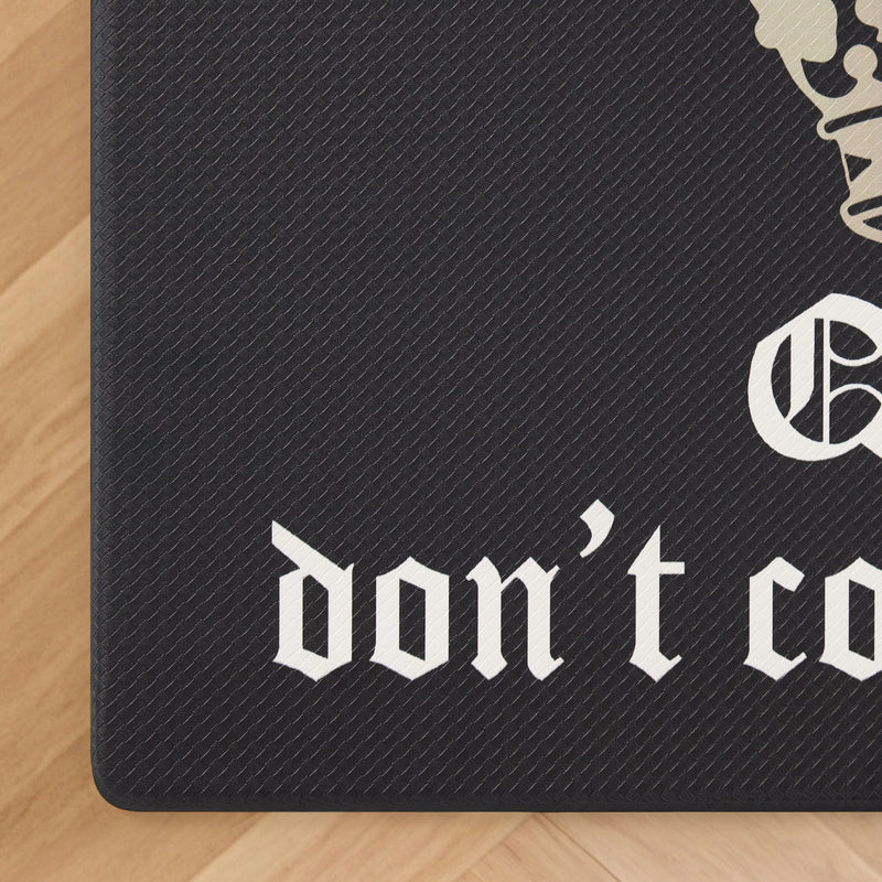 A black textured Kitchen Wellness Mat rests on a wooden floor, featuring the words “dont co” in a white Gothic-style font, with part of a decorative design and additional text partially visible at the top right corner. This cushioned anti-fatigue mat combines style and functionality—making it perfect as a kitchen floor mat or for adding comfort to any space.