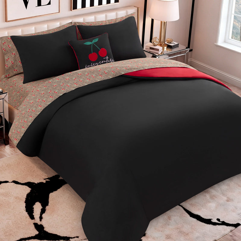 A neatly made bed features the Cherry Toss Cheetah Solid Reversible Comforter Bed in a Bag Bedding Set, showcasing black bedding with red accents and a cherry-themed pillow. The modern bedroom includes a lamp, books, and a window nearby, while small red floral details on the sheets and the plush bedding set add both coziness and designer-inspired style to the space.