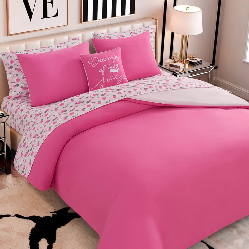 A neatly made bed features the Dreaming of You Solid Reversible Comforter Bed in a Bag Bedding Set in bright pink microfiber, paired with patterned sheets adorned with hearts and a pillow that reads “Dreaming of Juicy” in white script. The cozy bedroom is completed with a bedside lamp, artwork, and a patterned rug.