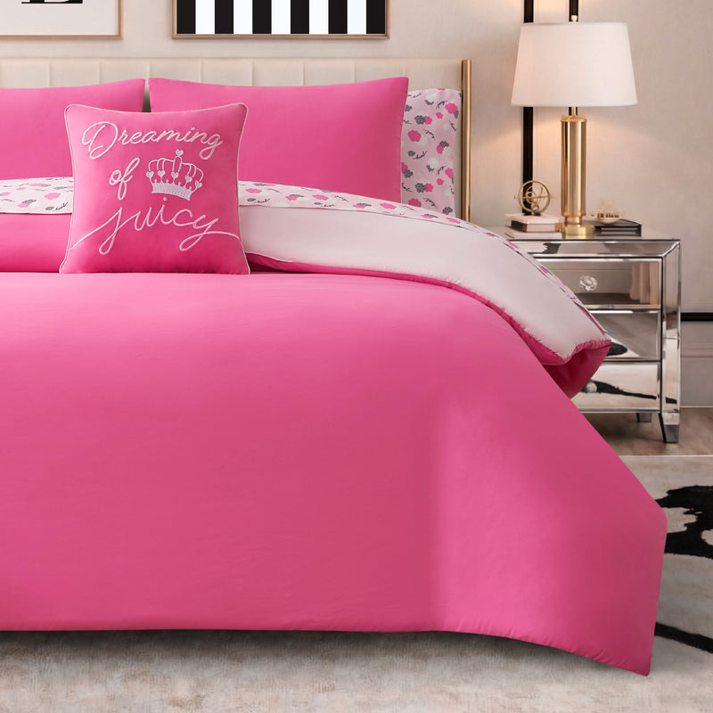 A neatly made bed is styled with the Dreaming of You Solid Reversible Comforter Bed in a Bag Bedding Set, featuring hot pink bedding, a decorative “Dreaming of Juicy” pillow, heart-patterned sheets, striped wall art, a lamp, and a mirrored nightstand topped with a clock and book. The reversible comforter provides versatile design options while the soft microfiber material promises cozy comfort throughout the night.
