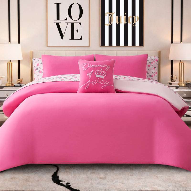A neatly made bed with a bright pink Dreaming of You Solid Reversible Comforter Bed in a Bag Bedding Set, a decorative pillow that says “Dreaming of Juicy,” and framed wall art reading “LOVE” and “Juicy” above the bed. Two lamps are on bedside tables.