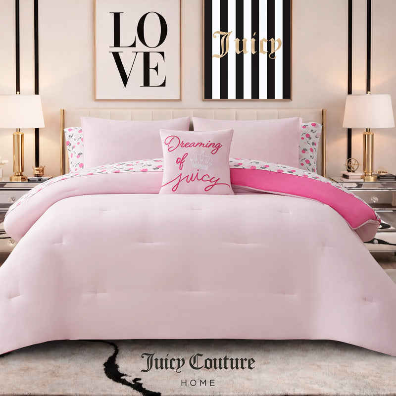 A pink Dreaming of You Solid Reversible Comforter Bed in a Bag Bedding Set from Juicy Couture is styled here, featuring a reversible comforter, a Dreaming of Juicy pillow, pink and white floral sheets, and framed LOVE and Juicy wall art above the bed. Two lamps rest on nightstands beside this chic Juicy Couture bedding ensemble.