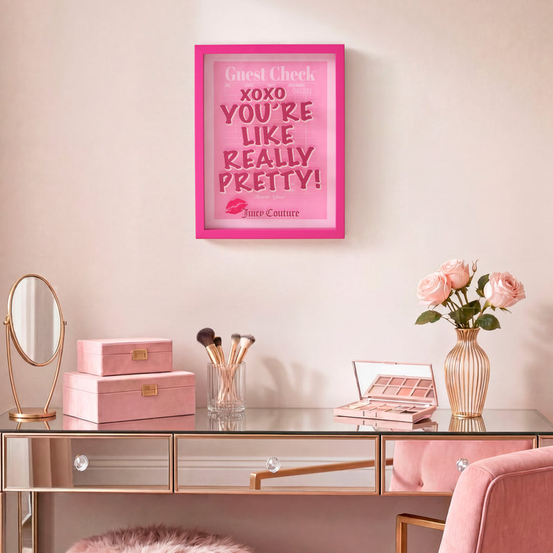 A vanity table with makeup, brushes, a mirror, pink velvet boxes, and a vase of pink roses sits beneath the Shadow Box Wall Art (12″ x 16″), a fashion-inspired framed pink sign that reads “YOU’RE LIKE REALLY PRETTY!” and adds a chic touch to the space.