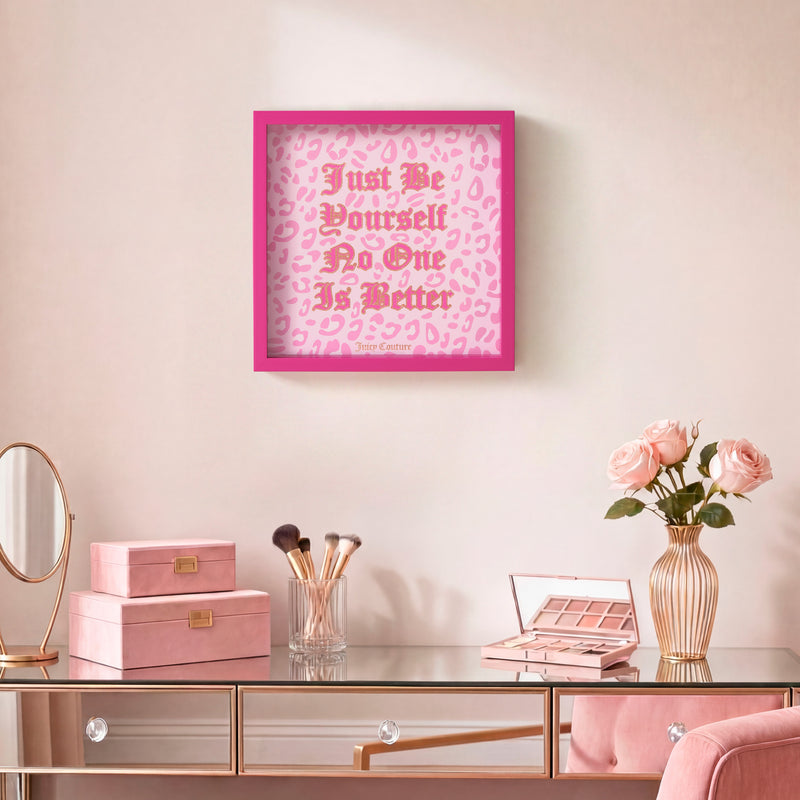 A Shadow Box Wall Art, 14″ x 14″, featuring a pink frame and the quote “Just Be Yourself, No One Is Better,” hangs above a vanity adorned with makeup, pink boxes, a mirror, and a gold vase of pink roses—adding a charming Juicy Couture-inspired accent to any glam space.